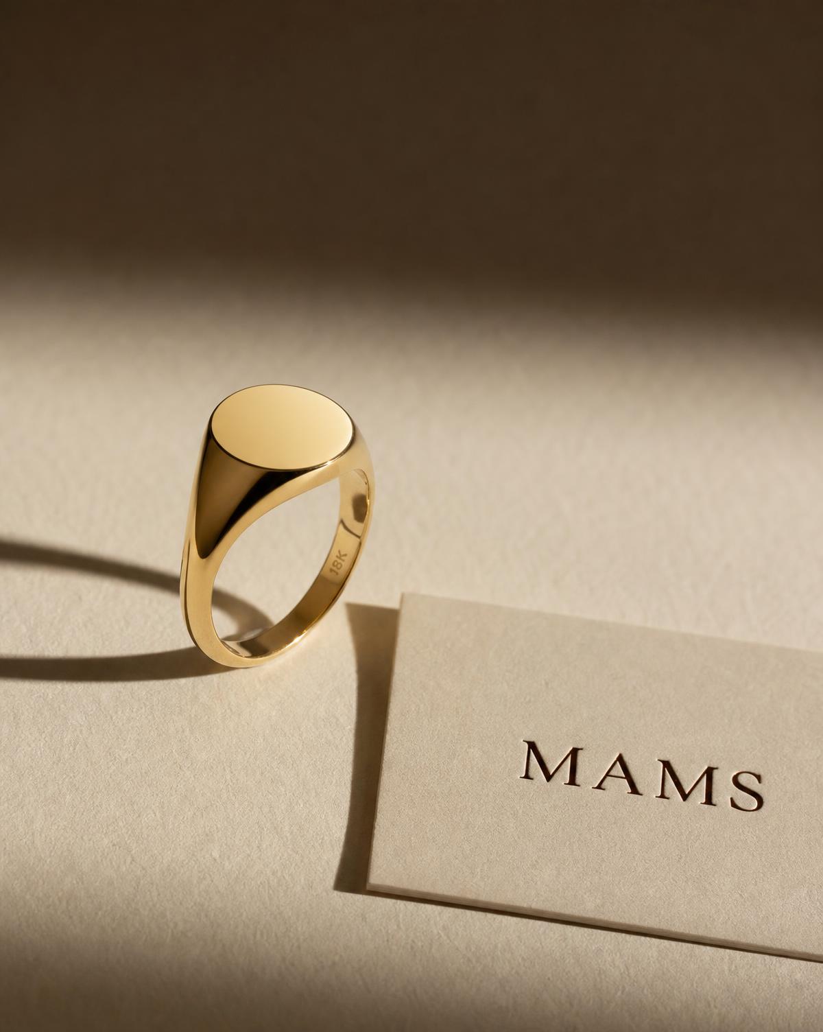 MAMS Legacy Signet Ring in solid 18K yellow gold, its plain polished face catching low light on ivory paper